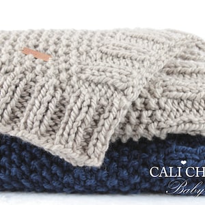Op de afbeelding: Twee gebreide dekens, een in lichtgrijs en de andere in donkerblauw, zijn op elkaar gevouwen. De bovenste deken heeft een klein bruin label met de tekst "CALI CHIC Baby" erop.