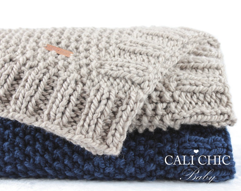 Op de afbeelding: Twee gebreide dekens, een in lichtgrijs en de andere in donkerblauw, zijn op elkaar gevouwen. De bovenste deken heeft een klein bruin label met de tekst "CALI CHIC Baby" erop.