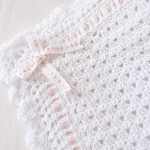 May include: A white crocheted blanket featuring a delicate openwork stitch pattern and a scalloped edge. A thin white ribbon with small pink accents is woven through the crochet work, tied into a small bow on the left side of the fabric.