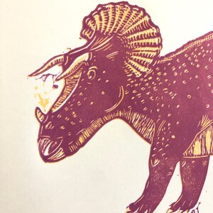 May include: A burgundy and yellow linocut print of a triceratops dinosaur. The dinosaur is facing left and has a large frill on its head.