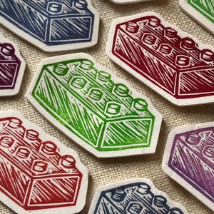 May include: Five colorful wooden block stamps, each depicting a different colored LEGO brick. The stamps are arranged in a semi-circle on a beige fabric background.