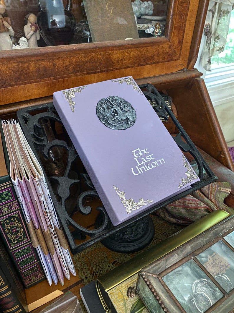 May include: A purple book with silver accents and a silver unicorn design on the cover. The title of the book is "The Last Unicorn".