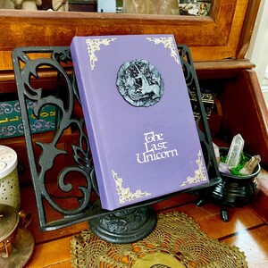 Another angle of The Last Unicorn book bound in light lavender purple leather, with silver corner filigree, title, and aged silver medieval crest affixed to the cover.