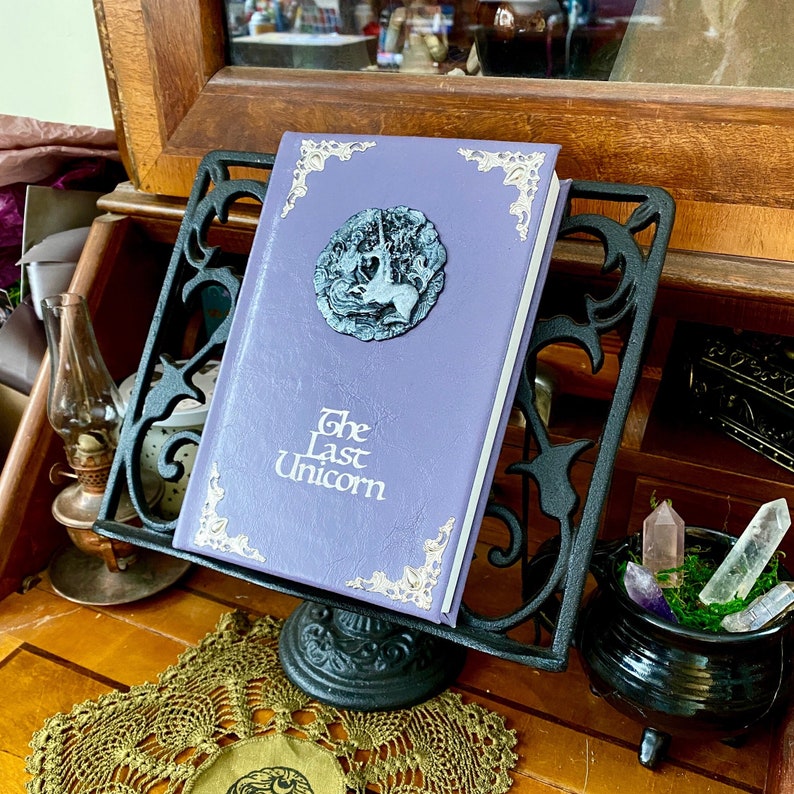 The Last Unicorn book bound in light lavender purple leather, with silver corner filigree, title, and aged silver medieval crest affixed to the cover.