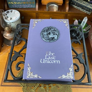 The Last Unicorn book bound in light lavender purple leather, with silver corner filigree, title, and aged silver medieval crest affixed to the cover.