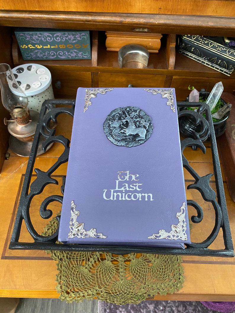 The Last Unicorn book bound in light lavender purple leather, with silver corner filigree, title, and aged silver medieval crest affixed to the cover.