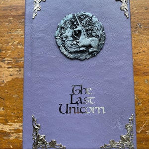 The Last Unicorn book bound in light lavender purple leather, with silver corner filigree, title, and aged silver medieval crest affixed to the cover placed on wooden desk