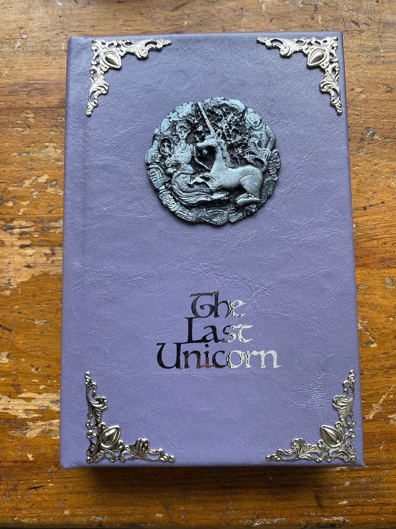 The Last Unicorn book bound in light lavender purple leather, with silver corner filigree, title, and aged silver medieval crest affixed to the cover placed on wooden desk