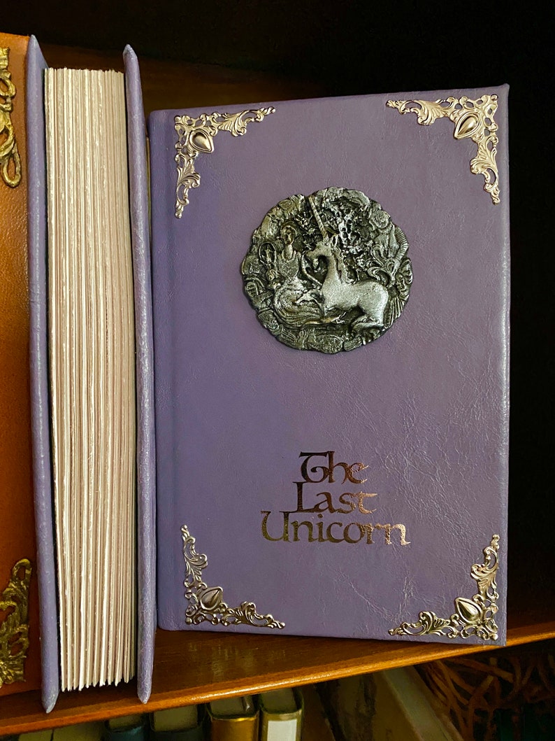 Photo shows the pages facing out to show deckled edges next to another copy of the Last Unicorn book in light purple leather with silver adornments