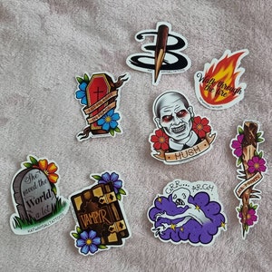May include: A set of ten colourful stickers featuring various pop culture and horror themed designs. The stickers include a coffin with the text "Let me rest in peace", a vampire book with the text "Vampyr", a tombstone with the text "She saved the world a lot", a monster with the text "Grr... Argh", a burning flame with the text "Walk through the fire", a stick figure with the text "Mush", a stick figure with the text "Mr. Pointy", a stick figure with the text "Katwhysalltattoos", and a stick figure with the text "Katwhysalltattoos".