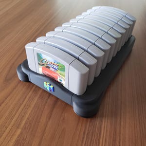 May include: A gray Nintendo 64 game cartridge holder with a row of gray game cartridges. The cartridges are labeled with game titles. The holder has the Nintendo 64 logo.