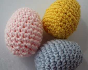 May include: Three handmade crochet eggs in pastel colors. One is light pink, one is yellow, and the other is light blue. The eggs are arranged in a triangular pattern against a white background. The crochet stitch creates a textured surface.