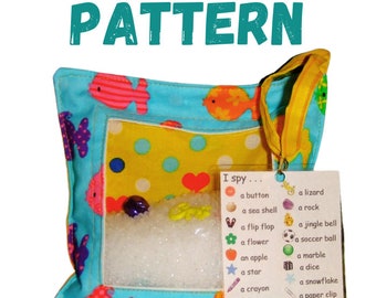 May include: A blue and yellow fabric pillow with a clear plastic window. The window shows white beads and colorful objects. A tag attached to the pillow says "I spy..." and lists various objects, such as a button, a sea shell, a flip flop, a flower, an apple, a star, a crayon, a mustache, a heart, a butterfly, a lizard, a rock, a jingle bell, a soccer ball, a marble, a dice, a snowflake, a paper clip, a dinosaur, and a gold ring.