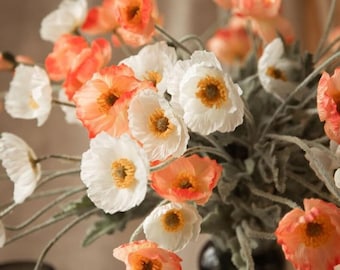 Puede incluir: Un ramo de flores de amapola artificiales con pétalos delicados en tonos blanco y naranja coral. Cada flor tiene un centro amarillo prominente y se sostiene sobre tallos verdes largos, finos y vellosos que se extienden en varias direcciones.