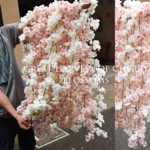以下が含まれることがあります： ピンクと白の人工桜の花が連なった長いガーランド。ガーランドはプラスチック製で、リアルな見た目です。ガーランドには「Great Harvest of Cherry Blossoms」とラベルが貼られています。