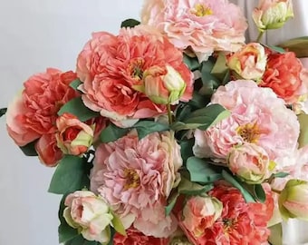 May include: A bouquet of artificial flowers featuring large, layered peony-style blooms in shades of coral, peach, and soft pink. The arrangement includes several fully opened blossoms, smaller partially closed buds, and green leaves.