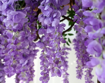 May include: Purple artificial wisteria flowers hanging from a branch. The flowers are in clusters and have a delicate, realistic appearance.