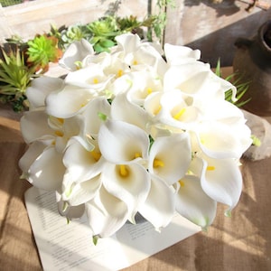 Puede incluir: Un denso ramo de calas artificiales con pétalos blancos lisos en forma de trompeta y espádices de color amarillo brillante. Las flores están dispuestas en un grupo redondeado, mostrando la forma curva y elegante de cada flor individual.