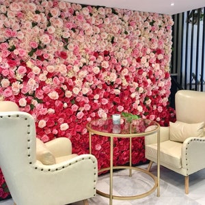 Könnte beinhalten: Zwei weiße Stühle mit Knopfpolsterung und ein goldfarbener Metalltisch mit Glasplatte stehen vor einer Wand, die mit künstlichen rosa und roten Rosen bedeckt ist.