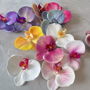 Pode incluir: Uma coleção de flores de orquídeas artificiais em várias cores, incluindo roxo, amarelo, rosa, branco e azul. As pétalas têm veias detalhadas e uma aparência realista. As flores estão dispostas sobre uma superfície cinza claro.