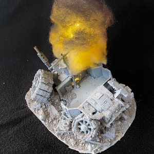 Könnte beinhalten: Graues Modell eines zerstörten Panzers auf einer strukturierten Basis. Das Modell zeigt Details wie Ketten, Räder und Komponenten. Ein gelb-grauer Wattebausch simuliert Rauch und Flammen, was eine Explosion andeutet.