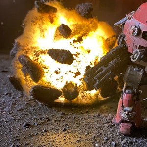 May include: A red and black robot figurine with a large gun, standing in front of a fiery explosion. The explosion is made of small, glowing rocks and debris.