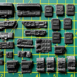 May include: A selection of small, dark grey plastic model components, such as dashboards, control panels, and electronic parts. The items are arranged on a green cutting mat with grid lines, indicating a hobby or model-making setting.
