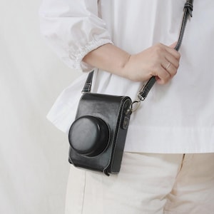 May include: Black leather camera case with a rounded lens cover and white stitching. The case has a shoulder strap and is designed to protect a camera. The case is shown hanging from a person's shoulder.
