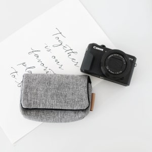 May include: A gray camera case with a black zipper and a brown leather pull tab. The case is sitting on a white surface next to a black Canon camera with a lens that says "Canon Zoom Lens 4.2-11.2 1:1.8-2.8 17X". The camera is on a white surface with the words "Together is our favorite place to go" written in black ink.