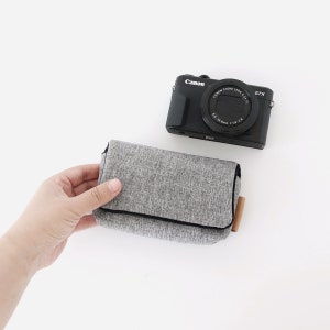 May include: A gray camera case with a black zipper and a brown leather pull tab. The case is made of a soft, textured fabric. A black Canon G7X camera is sitting on a white surface.