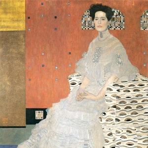 Op de afbeelding: Een vrouw in een witte jurk zit op een gepatroneerde stoel tegen een rode achtergrond. De stoel heeft een wit en zwart geometrisch patroon. De jurk van de vrouw is wit met een kanten overlay. Ze heeft donker haar en draagt een parelketting.