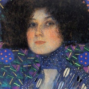 Op de afbeelding: Een vrouw in een blauwe jurk met een geometrisch patroon. De jurk heeft een hoge kraag en lange mouwen. Het haar van de vrouw is donker en krullend. De achtergrond is donkerblauw.