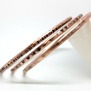 May include: Three copper bangles with varying textures. One bangle features a deeply hammered, vertical notch pattern, another has a repeating curved indentation design, and the third has a smooth, lightly distressed finish with a metallic sheen.