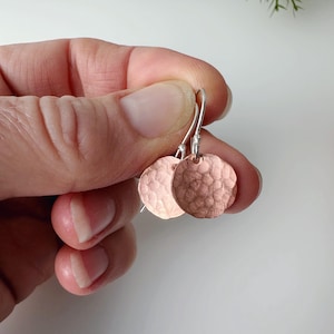 May include: A pair of copper-coloured, hammered-metal disc earrings with silver-coloured ear wires. The earrings are round and have a textured surface. The earrings are held in a hand against a white background.