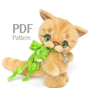 Könnte beinhalten: PDF-Muster für ein Plüschtier-Katze mit einer grünen Schleife und einem silbernen Anhänger. Die Katze ist hellbraun mit grünen Augen und weißen Schnurrhaaren.