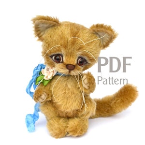 May include: A handmade, plush, tan-colored cat with large, expressive eyes and delicate whiskers. It has a blue ribbon with a flower around its neck. The image includes the text "PDF Pattern". This soft toy is a charming addition to any collection.