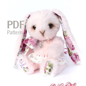 Puede incluir: Un conejito de peluche rosa con orejas de tela floral y una falda de encaje blanca. El conejito está sentado sobre un fondo blanco. Patrón PDF.