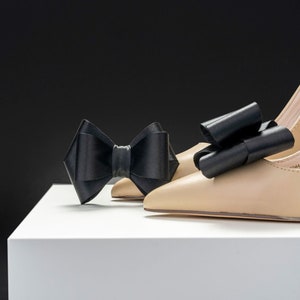 Pode incluir: Um par de sapatos de salto alto bege com la&ccedil;os de cetim preto nos dedos, sobre uma plataforma branca.