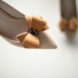 Könnte beinhalten: Ein Paar beigefarbene High-Heel-Schuhe mit einer braunen Schleife aus Wildleder an der Spitze. Die Schleife hat ein glitzerndes schwarzes Band in der Mitte.