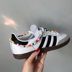 Peut inclure: Des baskets Adidas Samba en cuir blanc avec des bandes noires et une semelle en caoutchouc marron. Les chaussures ont une sangle de cheville perlée colorée avec de petits charmes de fleurs.