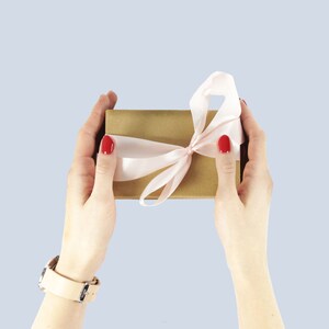 Pode incluir: Uma caixa de presente marrom com uma fita de cetim rosa amarrada em um la&ccedil;o. A caixa est&aacute; sendo segurada por duas m&atilde;os com esmalte de unha vermelho.