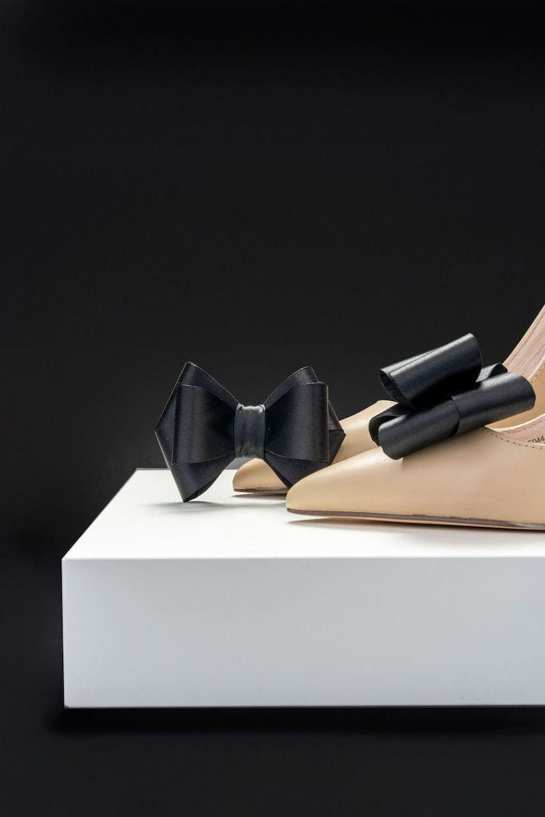 Pode incluir: Um par de sapatos de salto alto bege com la&ccedil;os de cetim preto nos dedos, sobre uma plataforma branca.