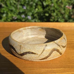 以下が含まれることがあります： ベージュ色の質感のある表面を持つ、浅い円形の陶器製のボウル。容器の外側には、手描きされた濃い茶色の波線が周囲を囲むように施されており、明るい斑点のある粘土の質感と対照的なコントラストを生み出しています。