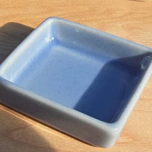 May include: A square ceramic dish with a light blue speckled glaze and rounded corners. The dish features a smooth, glossy finish with a deep interior basin and thick, raised walls, creating a simple, functional form for holding small items.