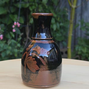 May include: A ceramic vase with a glossy black glaze on the upper portion and a matte brown finish on the lower base. The vase features a hand-painted, textured brown leaf design on the front, a narrow neck, and a wide, rounded body shape.