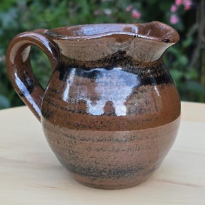 May include: A small, round ceramic jug with a glossy finish, featuring a wide handle and a curved pouring spout. The surface displays horizontal bands of earthy brown and dark black glaze, with a speckled texture throughout the body.