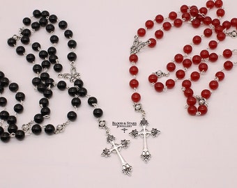 May include: Two beaded necklaces with silver cross pendants. One necklace has black beads and the other has red beads. The necklaces are on a white background.