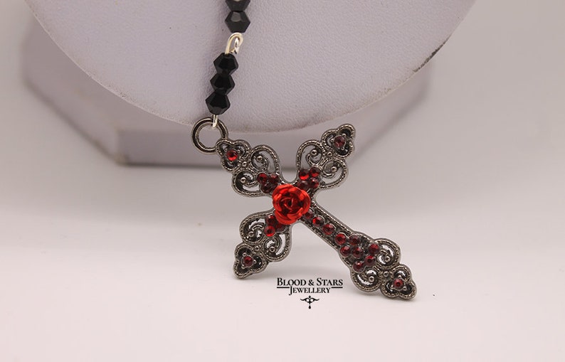 May include: A black beaded necklace with a silver filigree cross pendant adorned with red rhinestones and a red rose. The pendant features the text "Blood & Stars Jewellery".