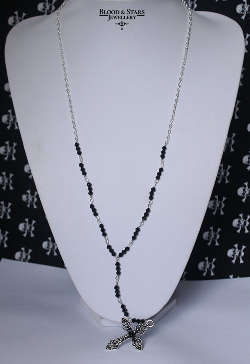 May include: A silver chain necklace with black beads and a silver filigree cross pendant. The necklace is displayed on a white mannequin bust against a black and white skull pattern background. The text "BLOOD & STARS JEWELLERY" is visible at the top of the image.