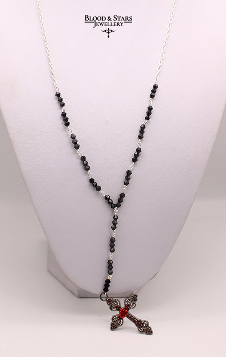 May include: A silver chain necklace with black beads and a silver cross pendant with red crystals. The cross has a detailed design and is accented with black crystals.